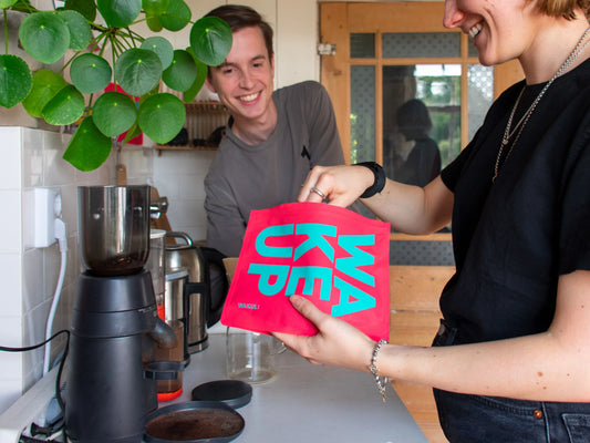 Koffie bewaren | Meisje met een roze Wakuli koffiezak in de hand in de keuken. Glimlachende jongen op de achtergrond.