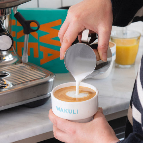Wakuli koffie proefpakket - Iemand giet opgeschuimde melk in een cappuccino in een keuken. Een zakje Wakuli koffie en een glas sinaasappelsap staan op de achtergrond.