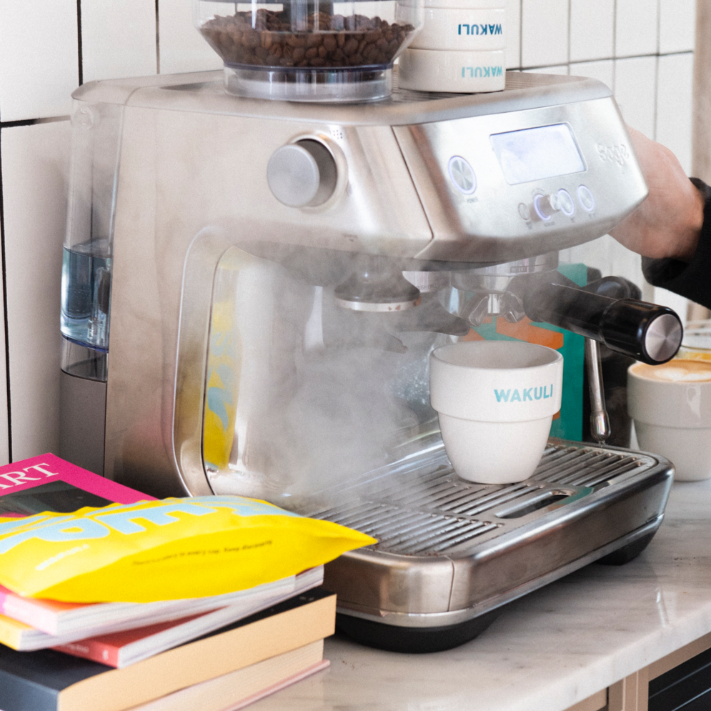 Discover Monthly Light Roast Koffiebonen - Er komt stoom uit een espressomachine. Wakuli zakjes en Wakuli koffiekopjes worden op en naast de espressomachine geplaatst.