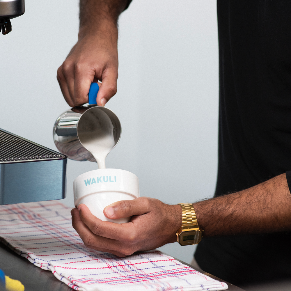 Koffiebonen voor latte - Iemand in een zwarte trui schenkt melk in een Wakuli koffiekopje. Links naast hem staat een koffiezetapparaat.