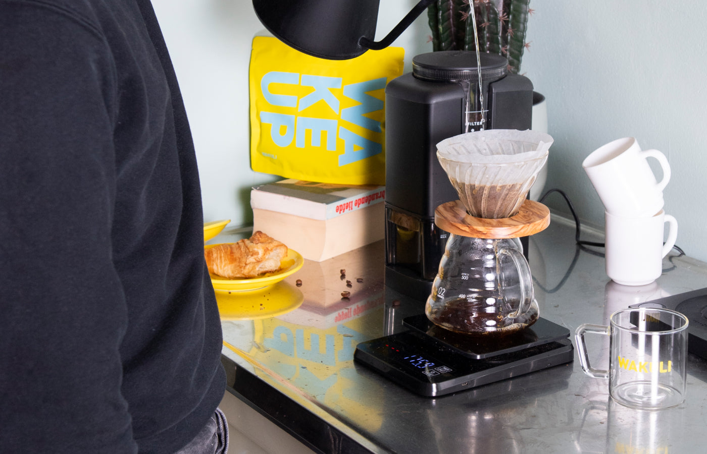 Koffie tools - Een jongen in een zwarte trui zet V60-koffie met een Hario dripper, een glazen kan en een gooseneck ketel. Croissant, handmaler, boeken en een geel Wakuli-zakje op de achtergrond.