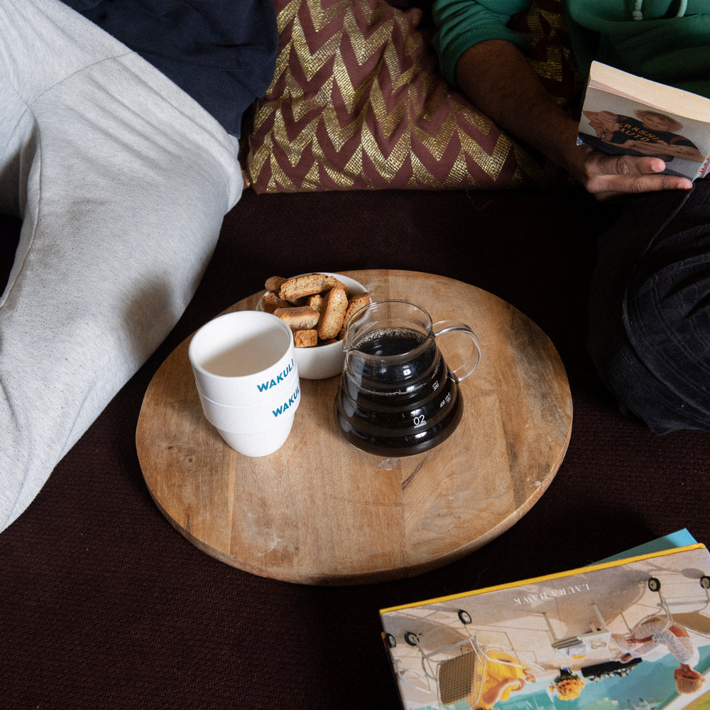 Filter koffiebonen proefpakket - Twee vrienden zitten op een bank. Eén leest een boek. Er staat een bruine ronde tafel met Wakuli koffiekopjes, kleine koekjes en een kan zwarte koffie erop.
