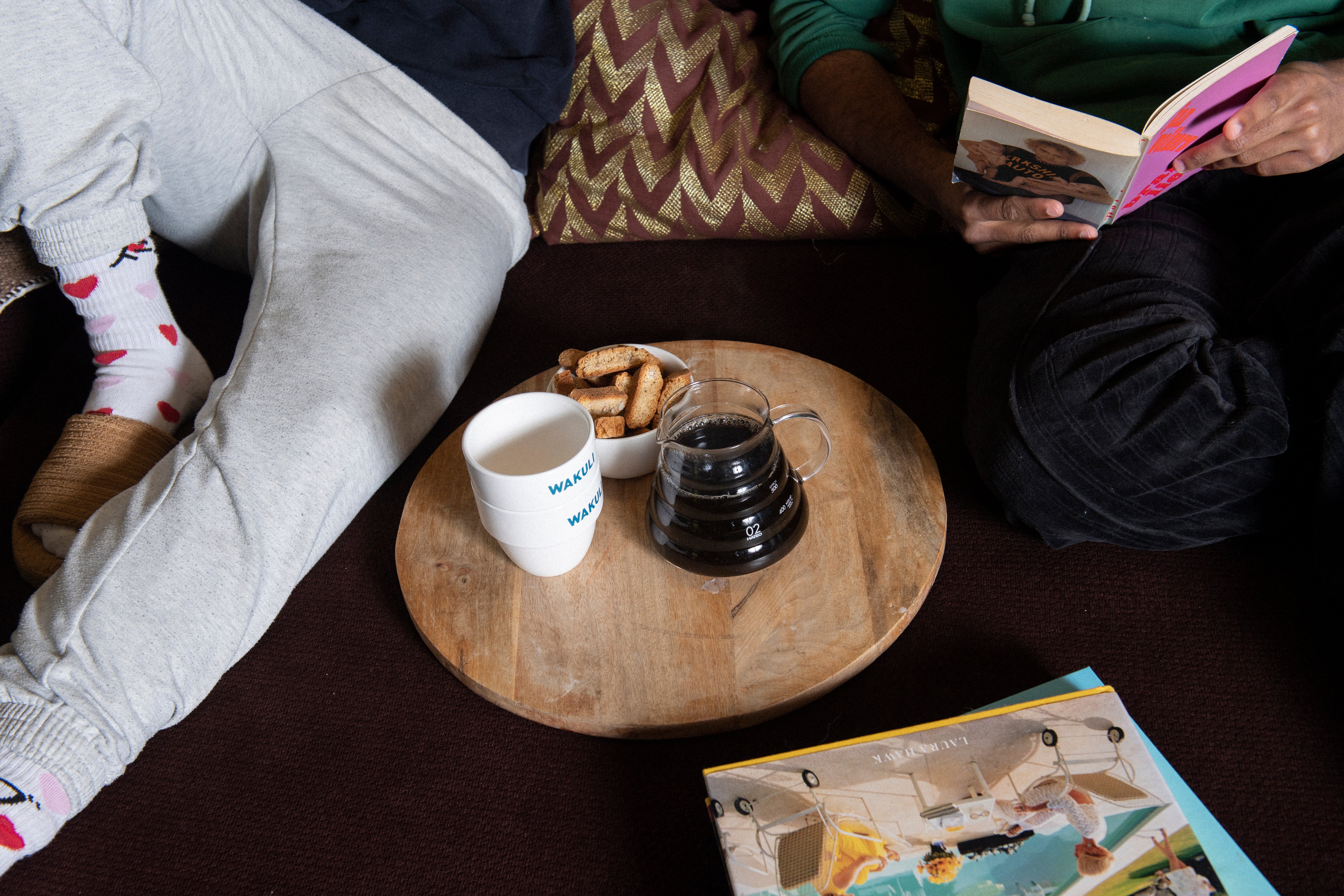 Filter koffiebonen proefpakket - Twee vrienden zitten op een bank. Eén leest een boek. Er staat een bruine ronde tafel met Wakuli koffiekopjes, kleine koekjes en een kan zwarte koffie erop.