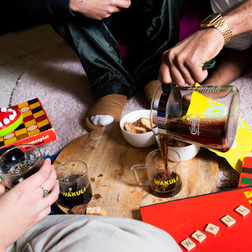 Verse koffiebonen proefpakket - Drie vrienden zitten rond een houten tafel. De een drinkt koffie uit een Wakuli-kopje, terwijl de ander zwarte koffie in een ander Wakuli-kopje schenkt. Op de tafel liggen verschillende spelletjes.