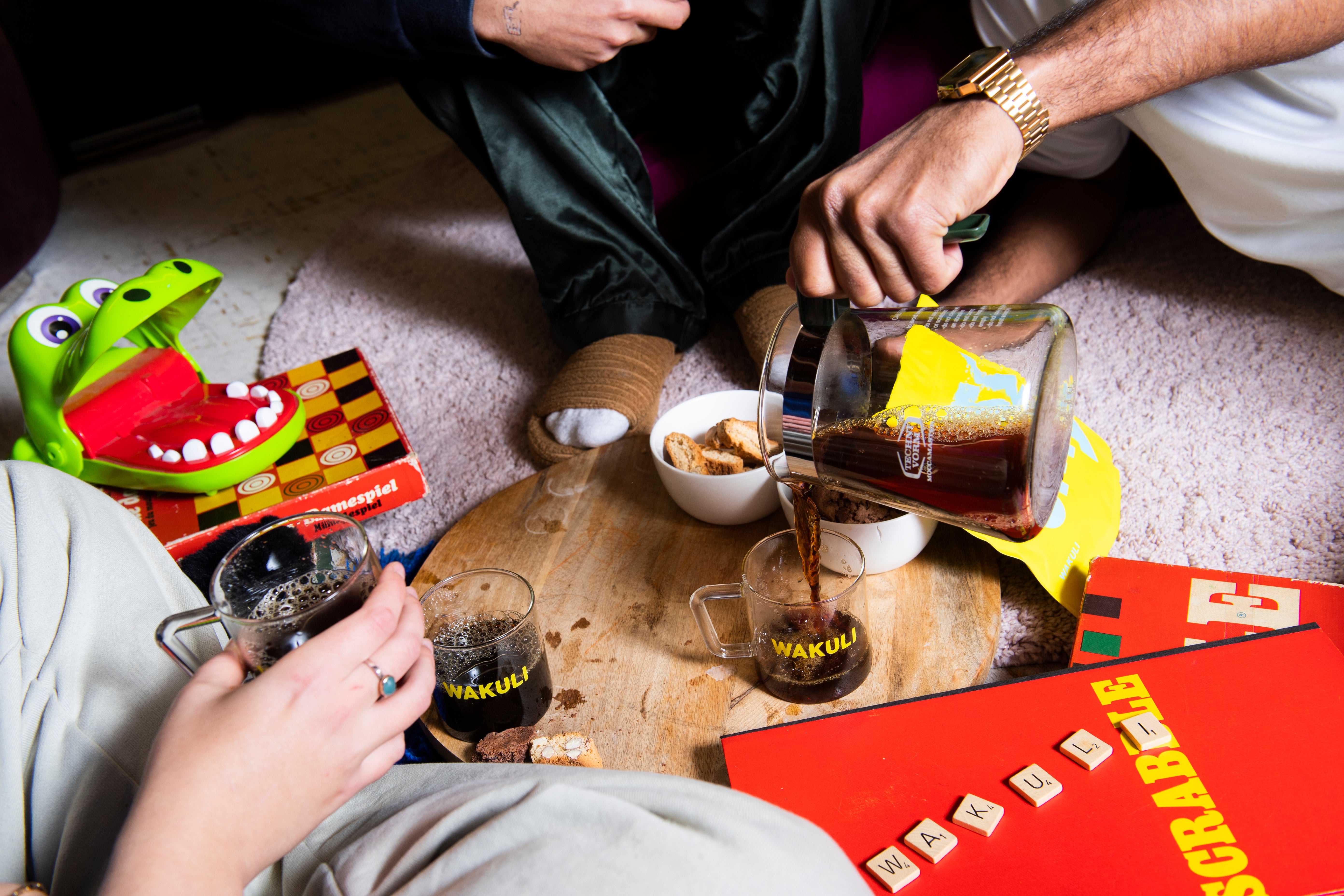 Verse koffiebonen proefpakket - Drie vrienden zitten rond een houten tafel. De een drinkt koffie uit een Wakuli-kopje, terwijl de ander zwarte koffie in een ander Wakuli-kopje schenkt. Op de tafel liggen verschillende spelletjes.
