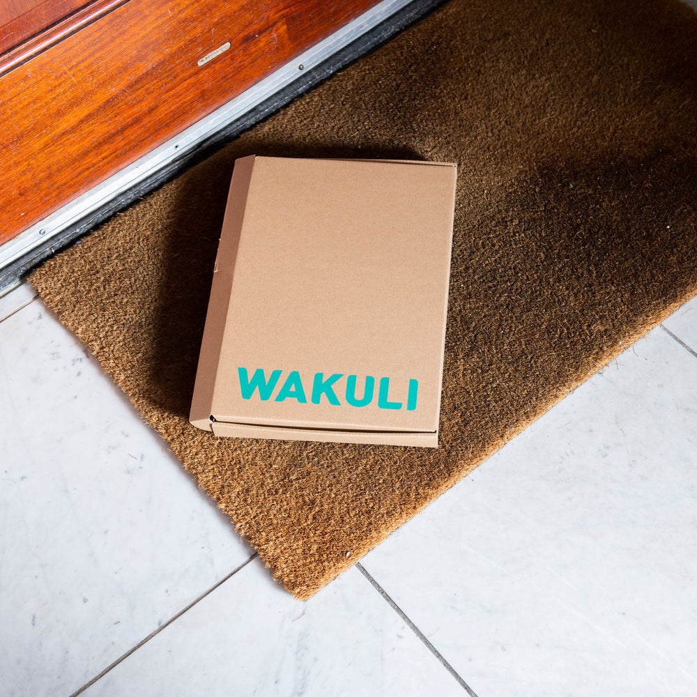 Koffiebonen voor latte - Een kartonnen doos met Wakuli erop ligt op een deurmat voor een bruine deur.