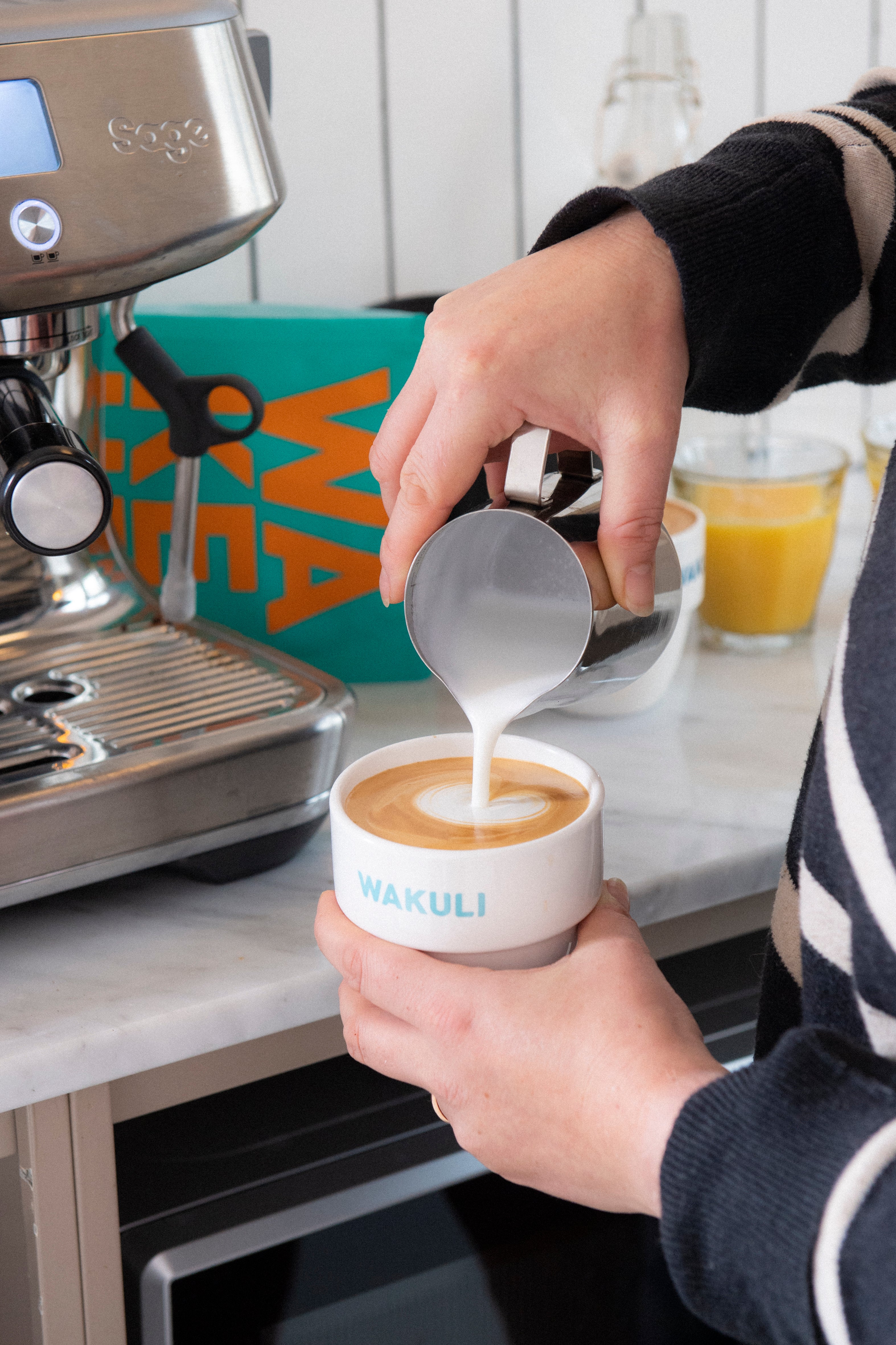 Koffiebonen voor latte - Iemand in een gestreepte trui schenkt melk in een latte in een Wakuli koffiebeker. Een espressomachine, een blauw Wakuli koffiezakje en een jus d'orange staan op de achtergrond.