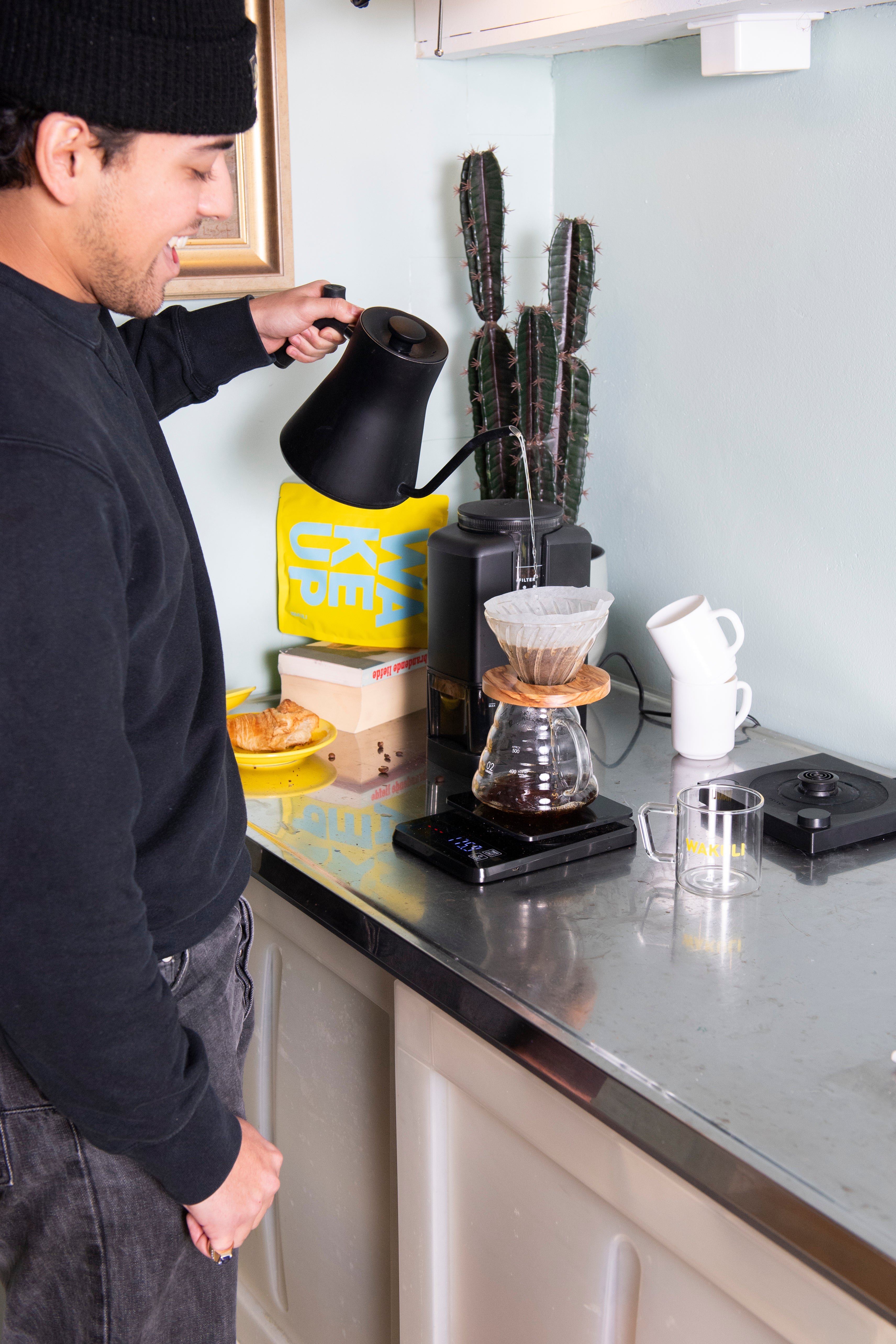 Filter koffiebonen proefpakket - Een jongeman met een zwarte hoed, spijkerbroek en trui schenkt water in een V60 koffieset. Er is een koffiemolen, een Wakuli koffiezakje, boek, een cactus en een croissant op een bord op de achtergrond.