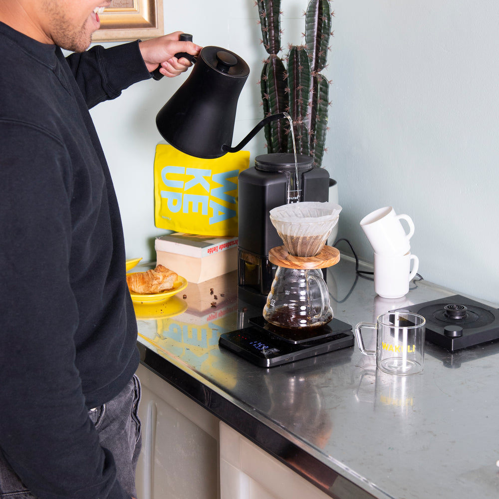 Filter koffiebonen proefpakket - Een jongeman met een zwarte hoed, spijkerbroek en trui schenkt water in een V60 koffieset. Er is een koffiemolen, een Wakuli koffiezakje, boek, een cactus en een croissant op een bord op de achtergrond.